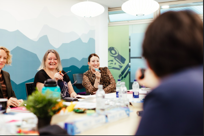 A soft focus image of some people on a training course. They are sat round a table, talking and laughing. The table is full of folders, pens, mugs, and training materials.
